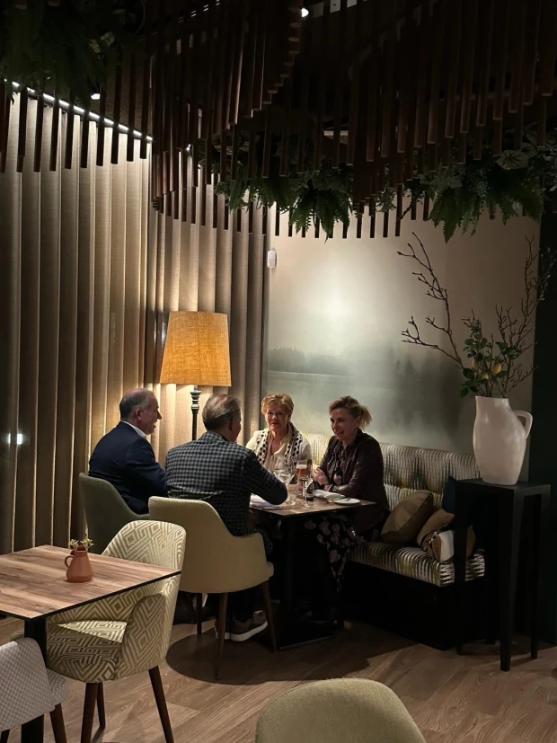 Vier gasten in gesprek aan een tafel in de brasserie, in het warme licht van een staande lamp, onder een plafond van houten lamellen en groen.