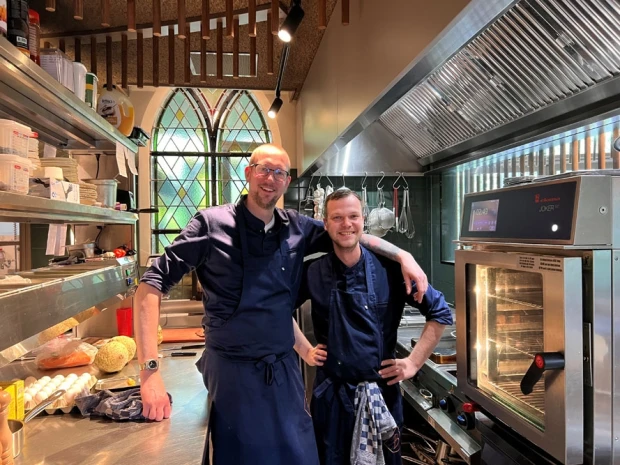 Twee koks in donkerblauwe schorten lachen in de keuken van Jacob van Bijstervelt, met een gotisch glas-in-loodraam achter hen.