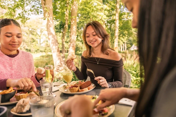 Drie vriendinnen eten samen aan een tafel in de tuin, onder hoge bomen in de zon, met hapjes, broodjes en glazen op tafel.