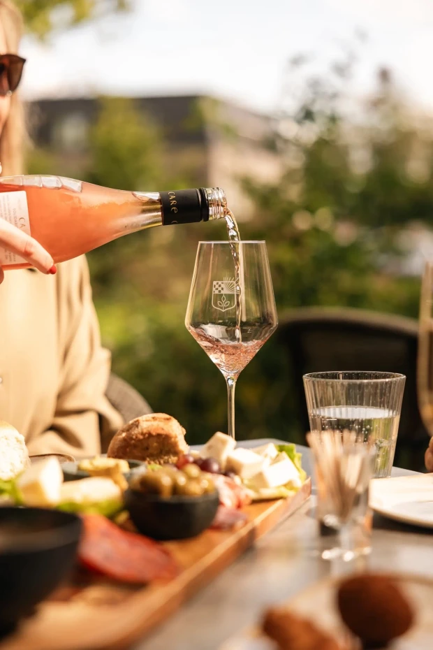 Roséwijn wordt ingeschonken in een wijnglas met het wapen van Jacob, op het terras, naast een plank met brood, kaas, olijven en worst.